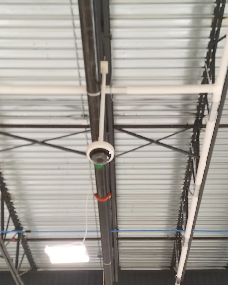 Axis high-bay dome camera suspended on a rigid EMT conduit drop from warehouse roof trusses at a distribution facility in Bolton, ON.