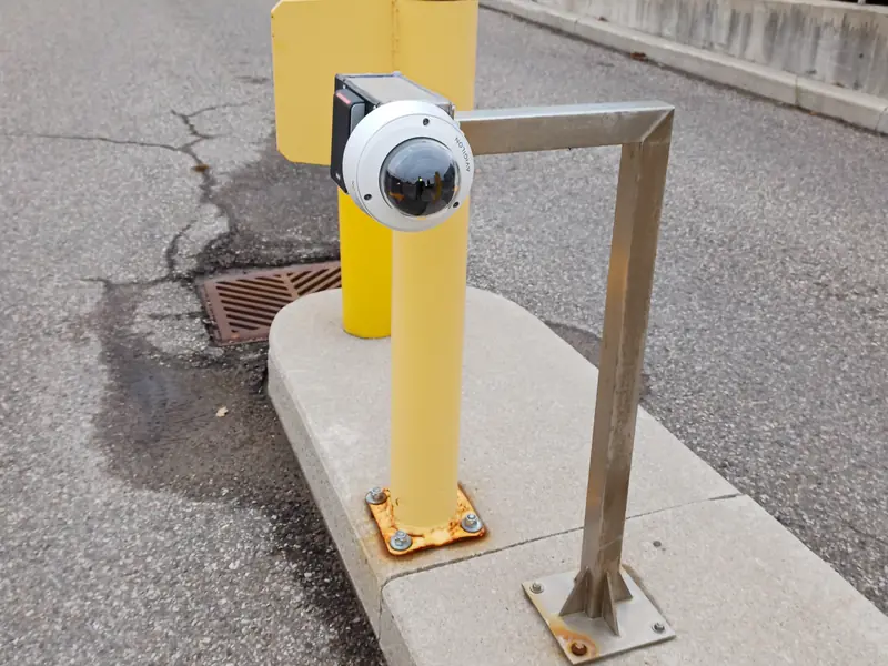 Axis license plate recognition (LPR) and vehicle lane camera mounted on a custom steel arm bolted to a yellow bollard at a 150,000 sq ft Vaughan, ON warehouse.