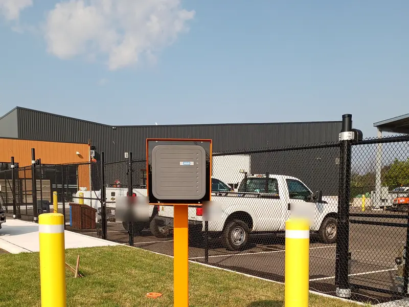 HID long-range UHF vehicle gate reader mounted on a high-visibility orange pedestal between steel bollards at a fenced logistics yard in Vaughan, ON.